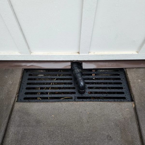 Drain grate positioned beneath a white wall, with a black pipe visible